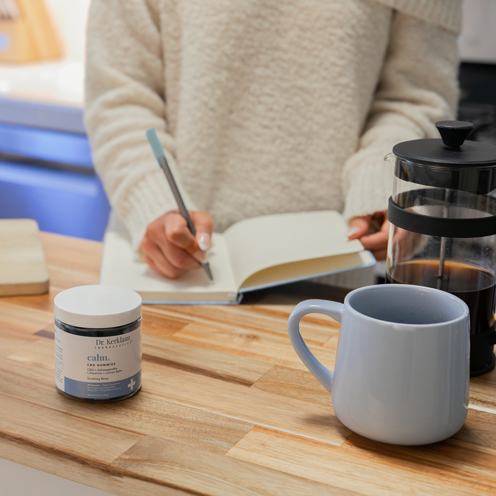 Jar of Dr. Kerklaan Calm CBD Gummies on kitchen counter beside coffee and journal, as a woman writes during her calming morning routine.