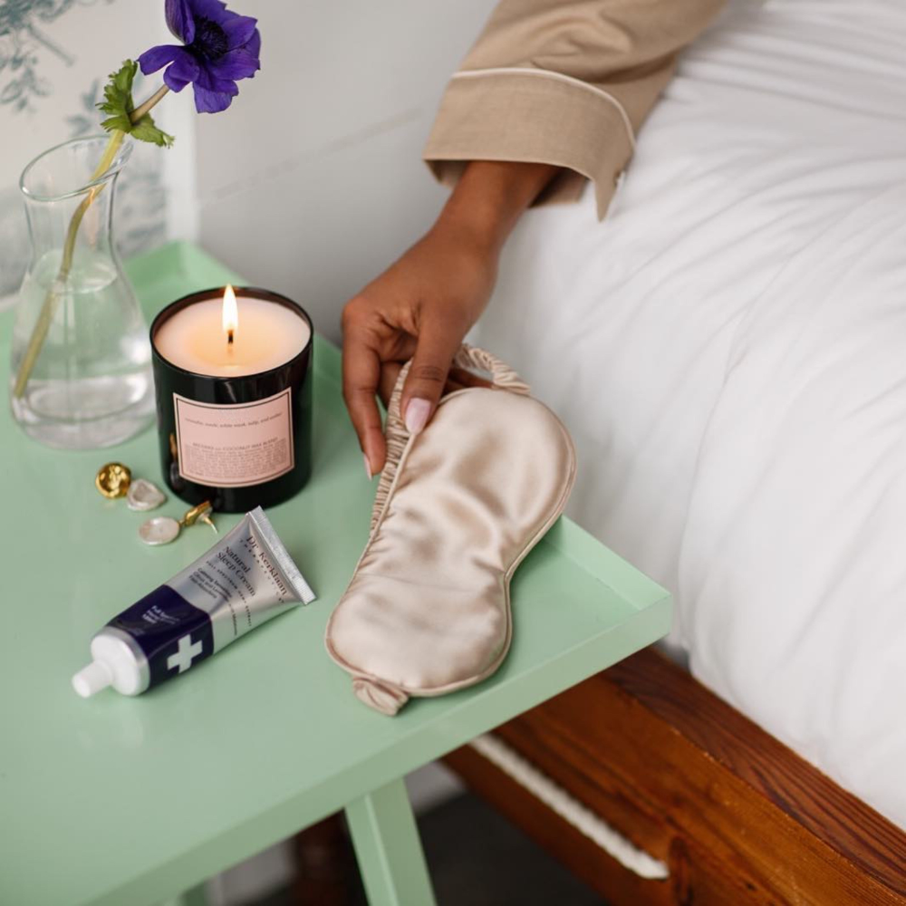 Natural CBD Sleep Cream placed on a bedside table with a candle, silk sleep mask, and jewelry, creating a relaxing nighttime setting.